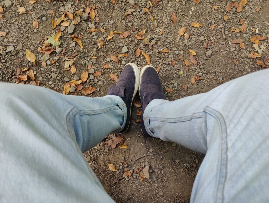 Downward view of my legs as I rest on a bench during a fall hike, with the leaf-strewn gravel path under my feet.